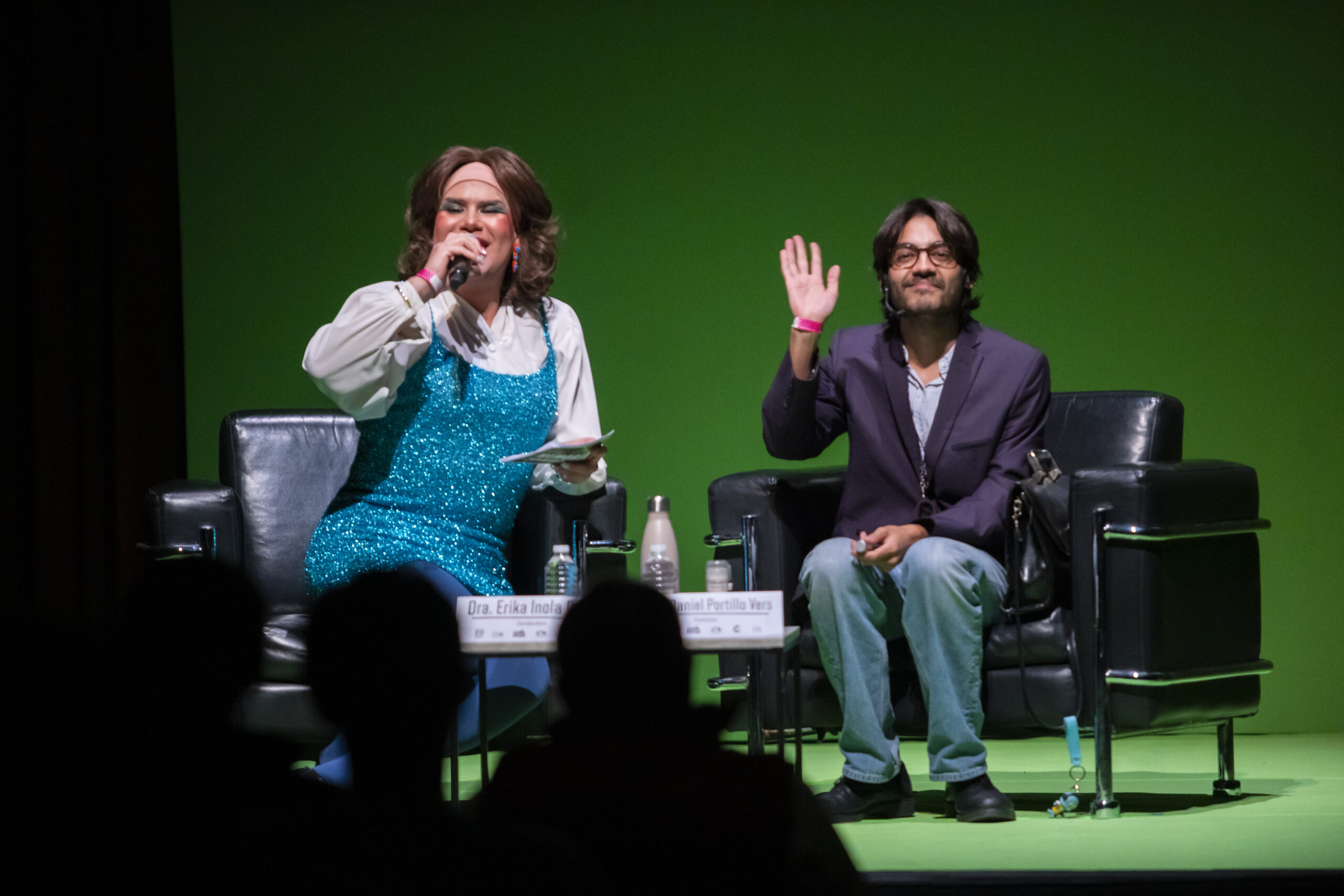 Una académica en vestido de lentejuelas y un desgarbado traductor saludan a la audiencia, sentadxs en dos sillones negros dentro de un espacio completamente verde.