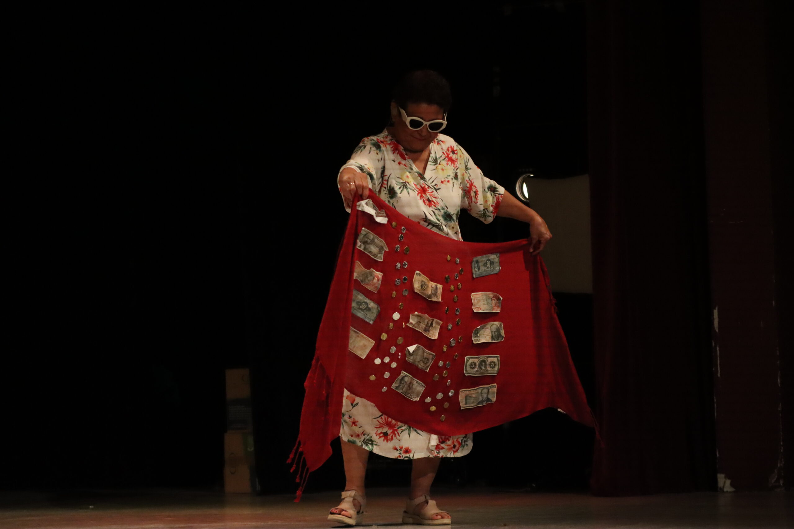 Una mujer mayor con un vestido floreado y lentes de sol muestra billetes y monedas de colección sobre una tela roja.